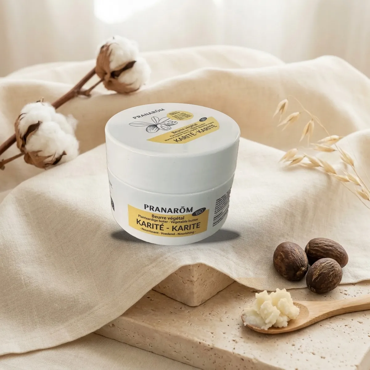 Le pot de beurre de karité bio et brut de Pranarōm, fermé, posé sur un linge crème à côté de noix de karité