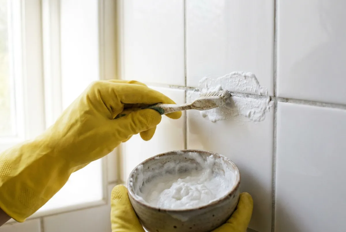 Une main gantée brosse la pâte de bicarbonate sur un joint gris entre deux carreaux blancs, un bol de pâte à la main